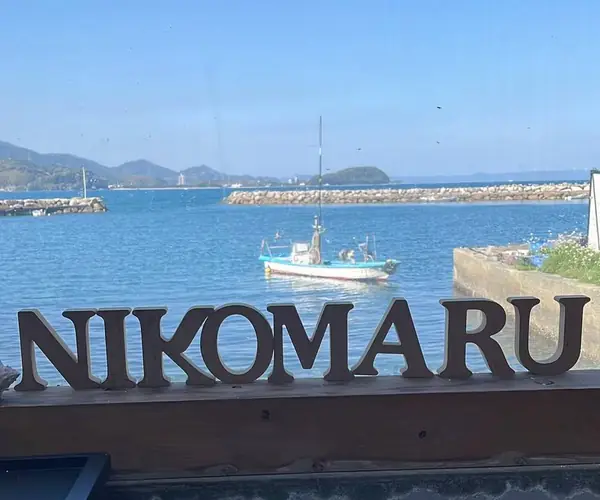 窓の外の青い海に浮かぶ漁船。手前に NIKOMARU の木の文字